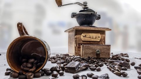 Wooden coffee grinder with beans and chocolate on a rustic table setting.