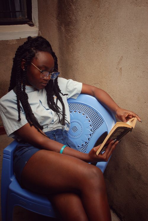 Free A young woman sits in a blue chair, engrossed in a book indoors. Casual and relaxed atmosphere. Stock Photo