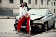 一位时尚女士自信地在一辆城区受损的汽车旁摆出姿势，散发着现代风格与大胆气息