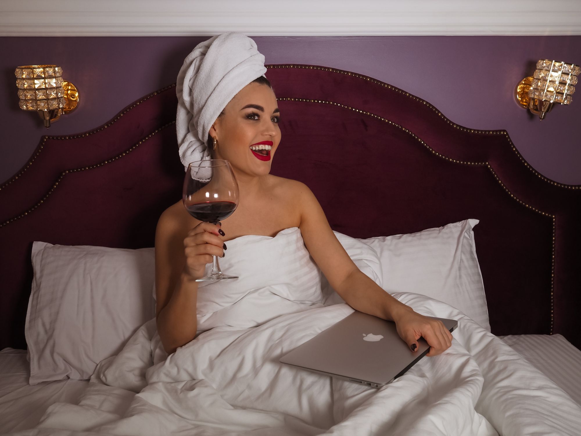 Smiling woman in bed with wine glass and laptop, enjoying a cozy evening indoors.