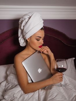 Woman sitting in bed with a towel head wrap, holding a laptop and red wine glass.