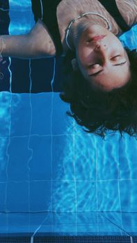 A woman with eyes closed floats serenely in a clear blue swimming pool under sunlight.