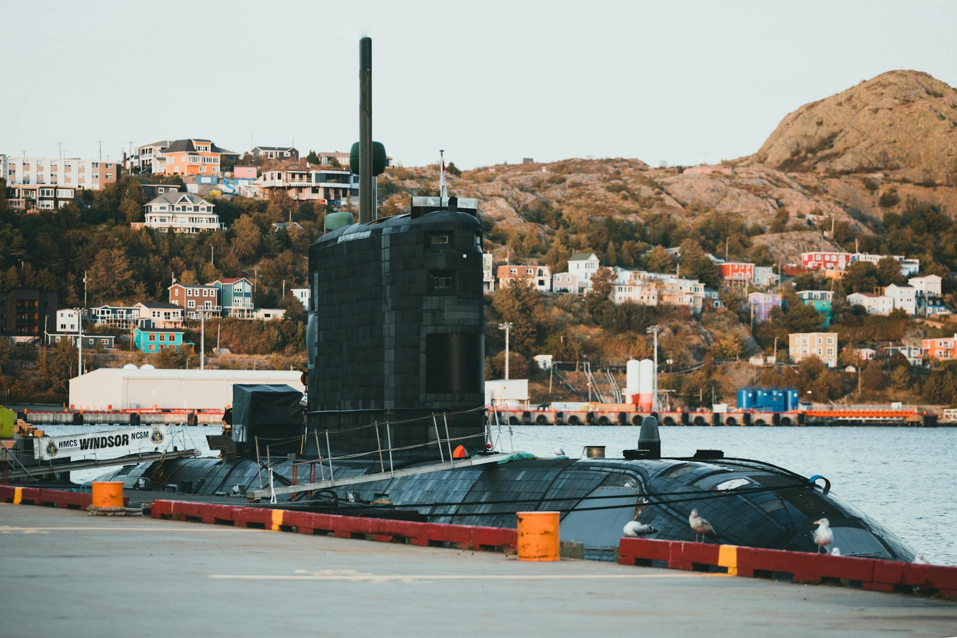 Submarine docked at a colorful hillside harbor with urban scenery.