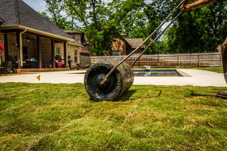 A worker rolling a lawn in a suburban backyard near a pool on a sunny day.