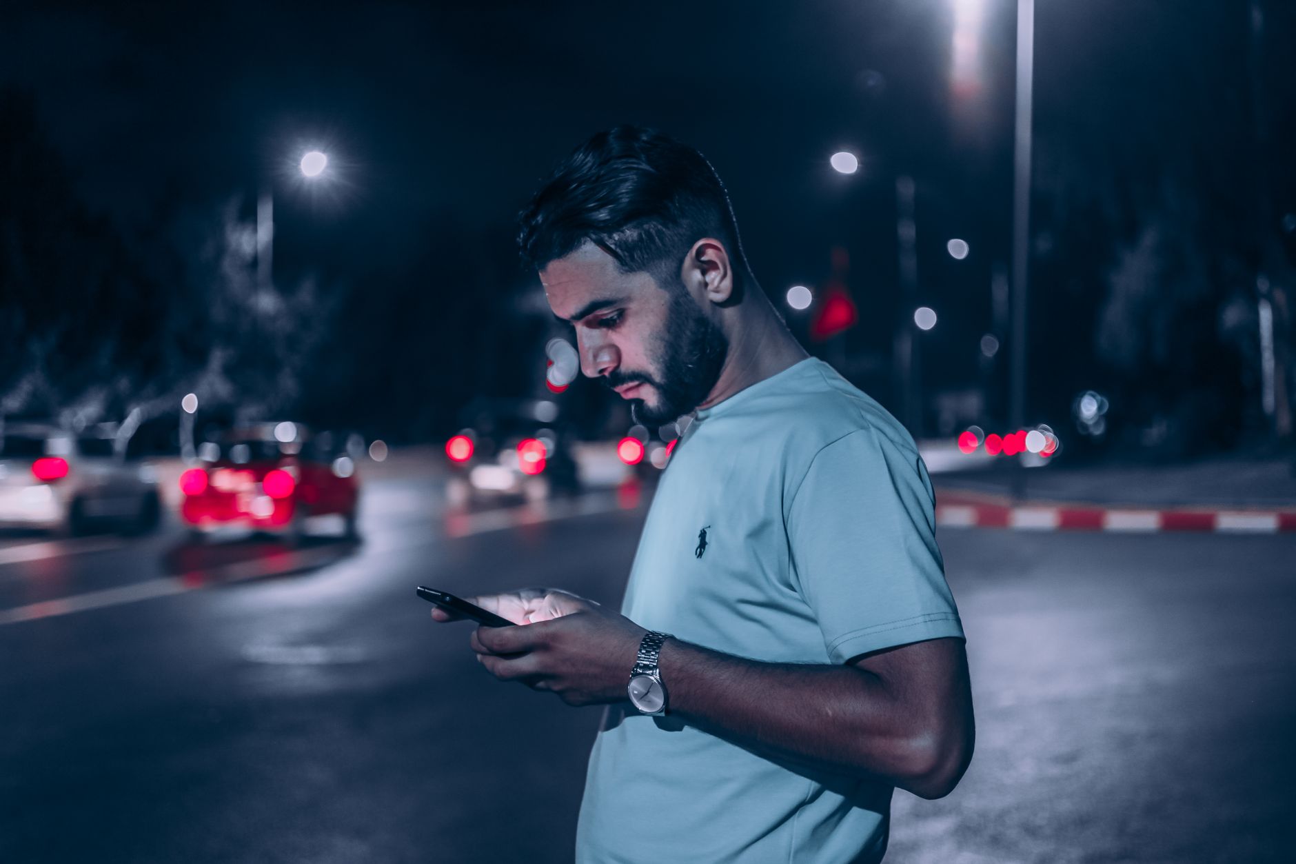 A man in a white shirt checks his phone at night on a busy city street, lit by shopfronts.