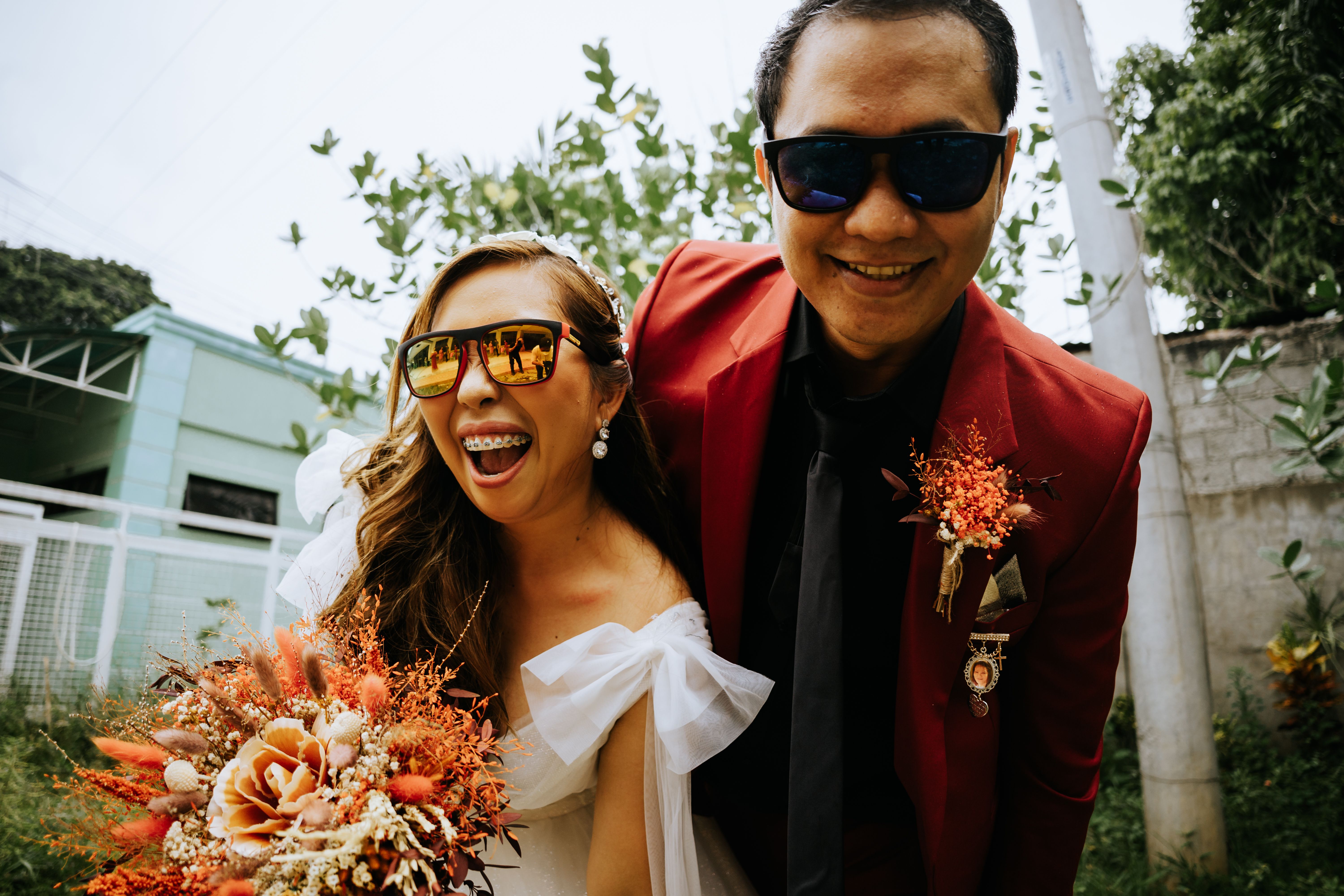 Happy couple in sunglasses smiling at their wedding with colorful bouquets.