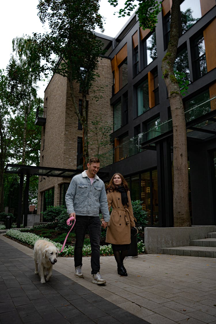 A Man In Denim Jacket And A Woman In Brown Coat Walking A Dog