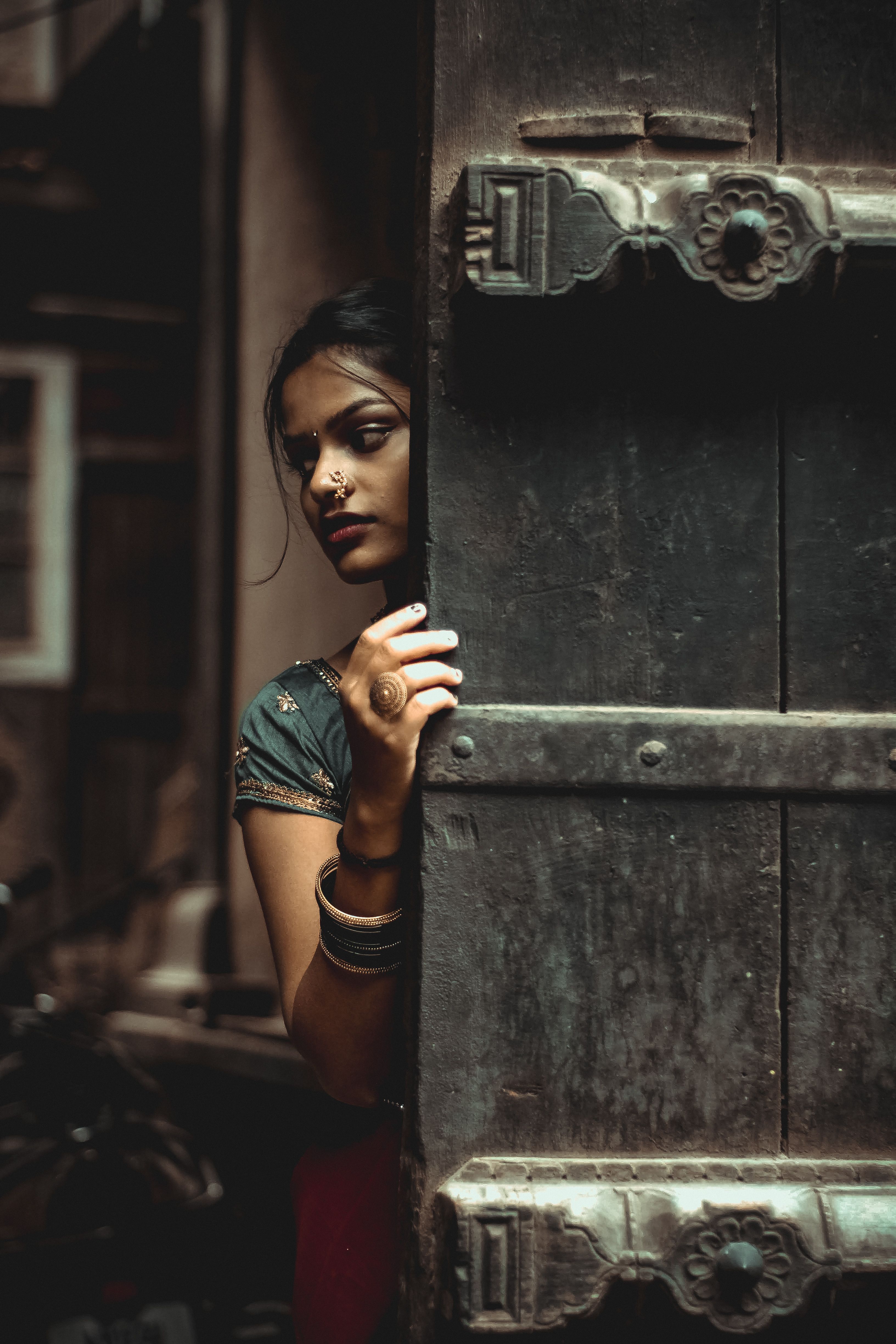 A woman with a nose piercing peeks behind a vintage wooden door, wearing bracelets.