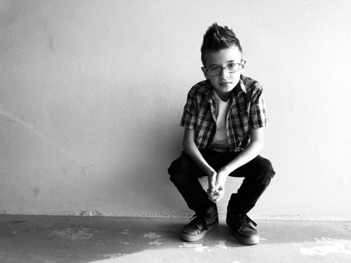 Free A stylish young boy squatting against a wall in black and white, wearing glasses. Stock Photo