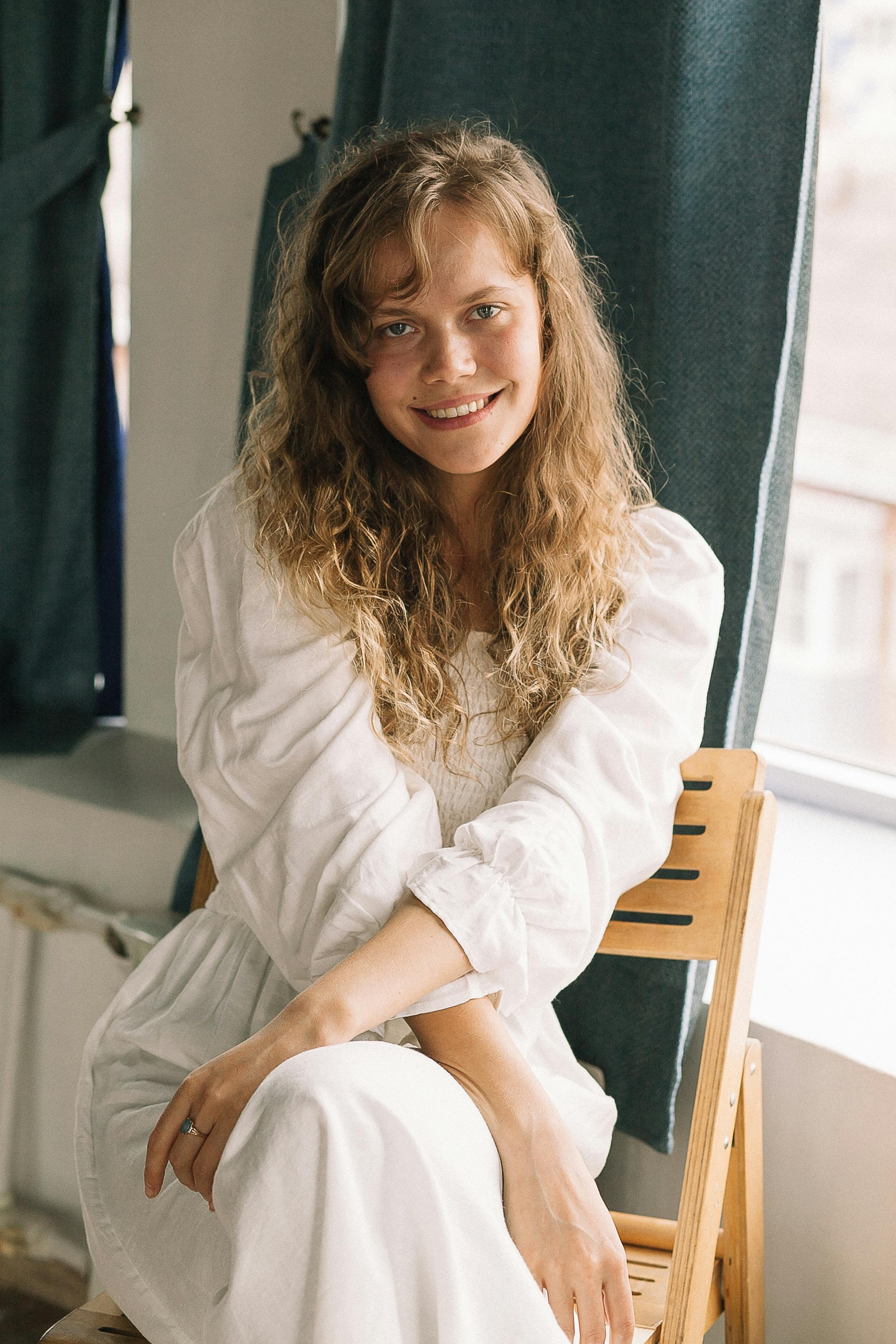 Smiling woman in a white dress sitting indoors, sunlight streaming through windows.