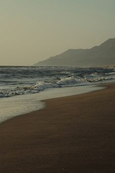 Serene sandy beach with gentle waves and distant mountains at sunset.