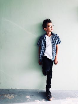 一个穿着格子衬衫的时尚小男孩靠在浅色墙壁上摆姿势，散发着自信