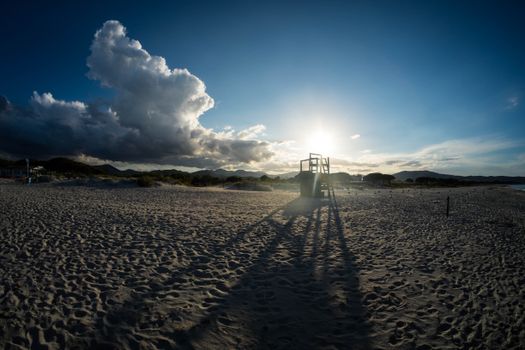 Stunning beach landscape at sunset with dramatic clouds and lifeguard tower casting long shadows on the sand.