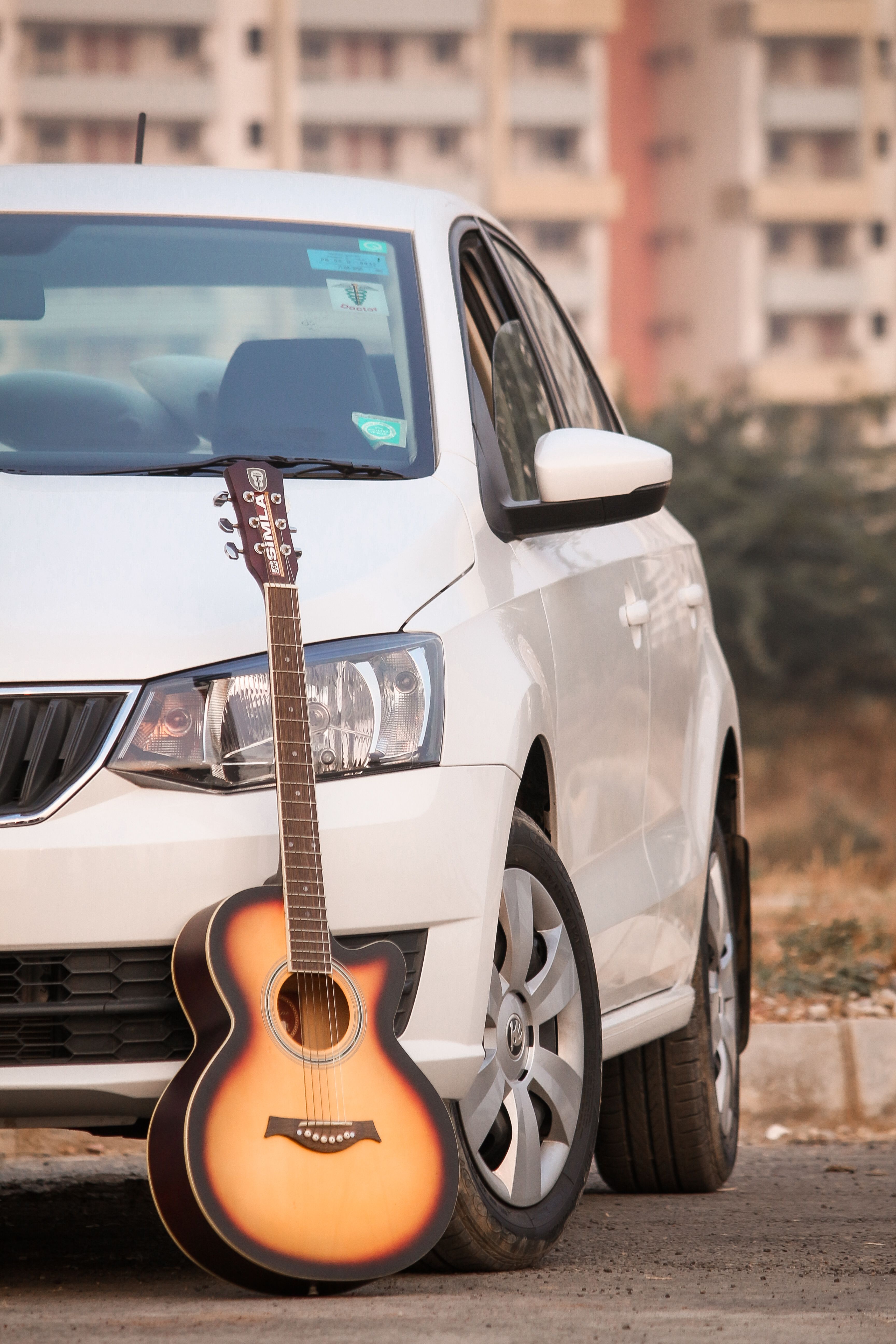 A classic acoustic guitar propped against a parked white car in an urban setting.