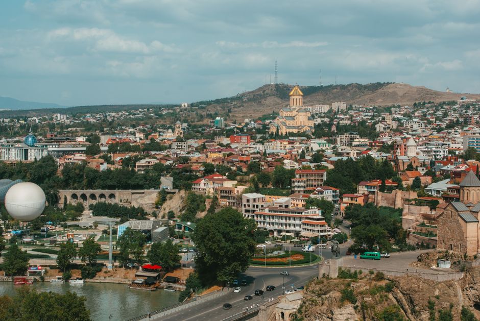 A panoramic aerial view of Tbilisi featuring the Holy Trinity Cathedral and surrounding urban landscape.