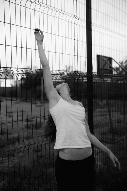 Artistic black and white photo of a woman stretching against a fence outdoors.