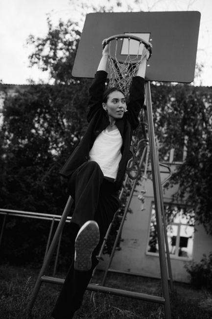 Black and white photo of a woman hanging on a basketball hoop, showcasing playful energy.