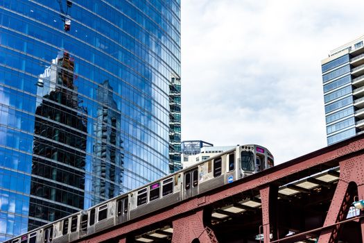 A Chicago train passes a modern glass skyscraper reflecting the cityscape.