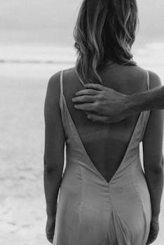 A woman in a backless dress stands on a beach with a hand gently resting on her shoulder.