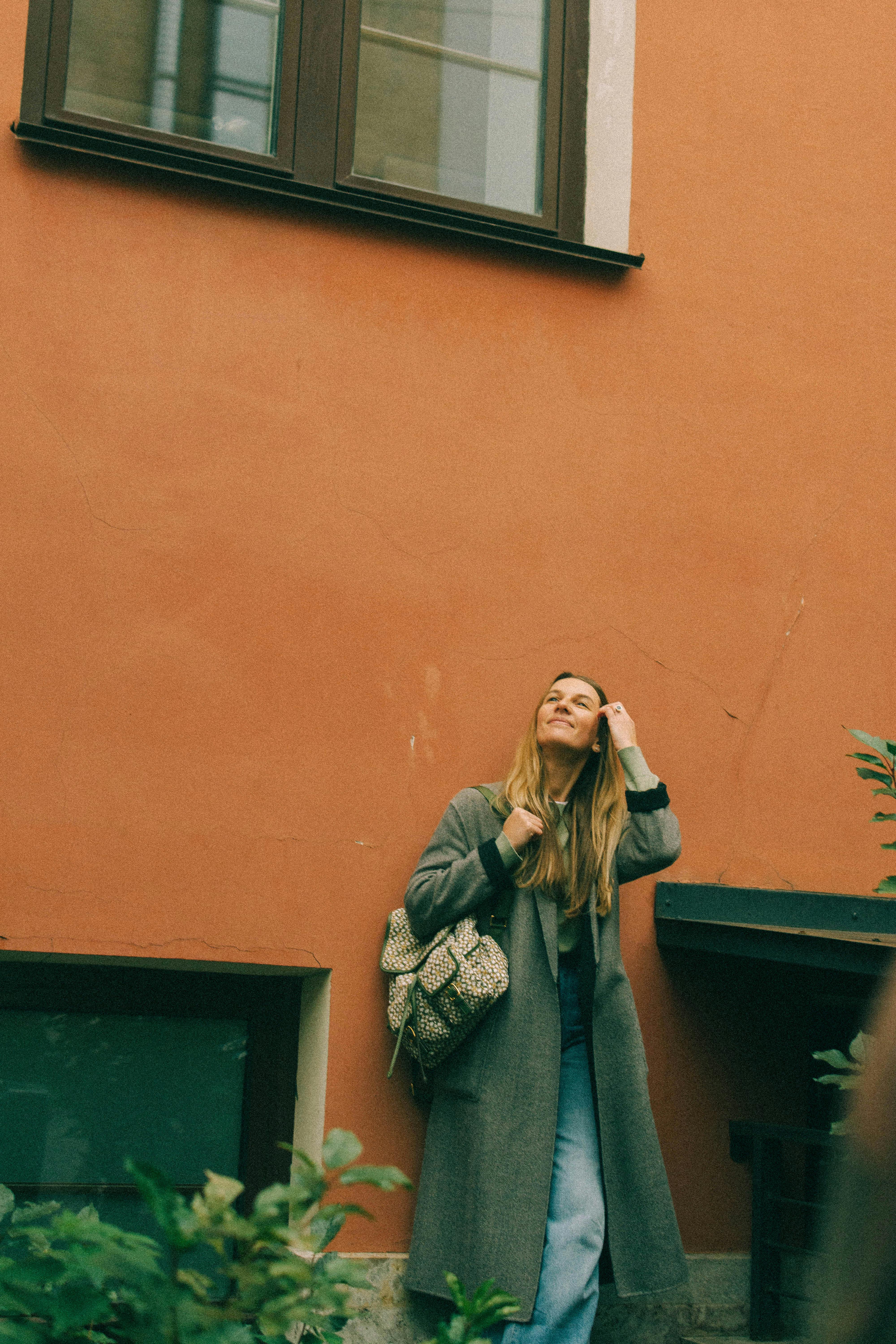 Caucasian woman in a coat with a backpack, leaning against an orange wall outdoors.