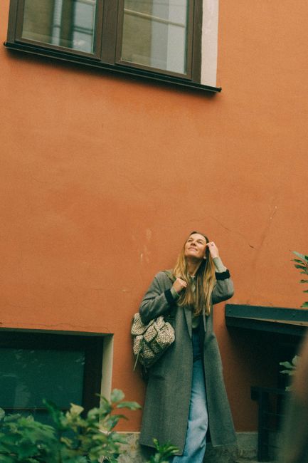 Caucasian woman in a coat with a backpack, leaning against an orange wall outdoors.