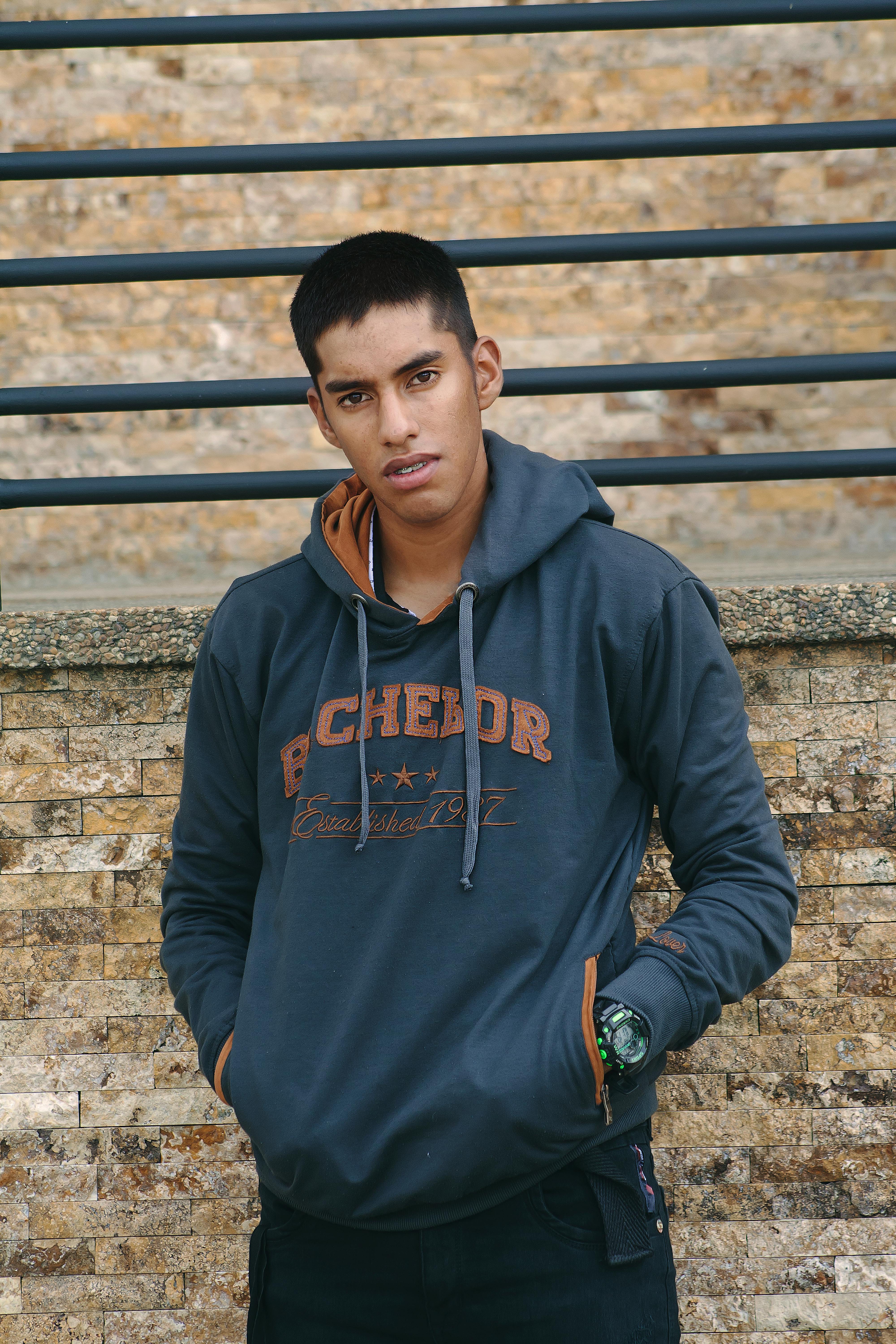 Portrait of a young man in a hoodie with hands in pockets, leaning against a brick wall outdoors.