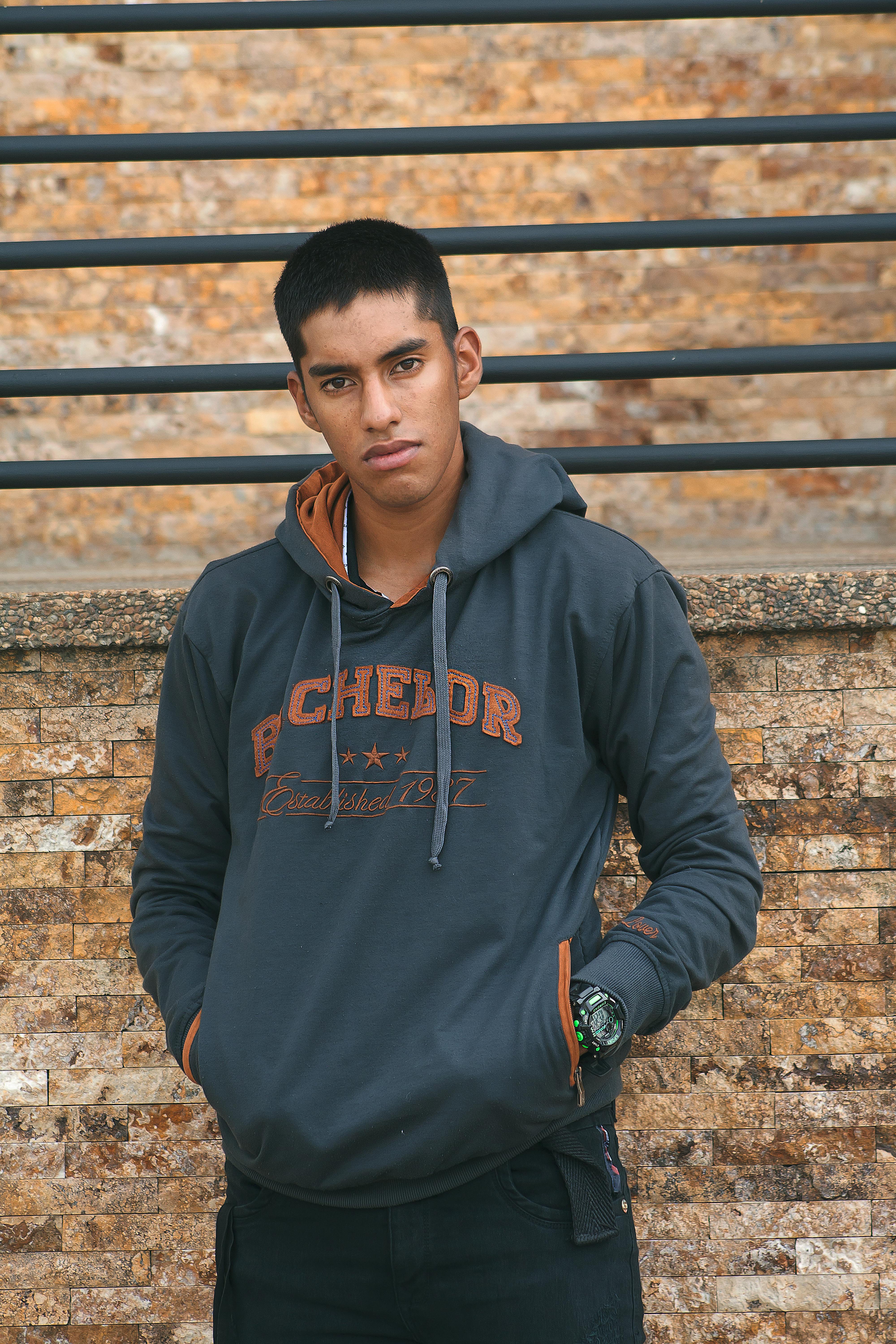 A young man in a hoodie stands pensively against a brick wall outdoors.