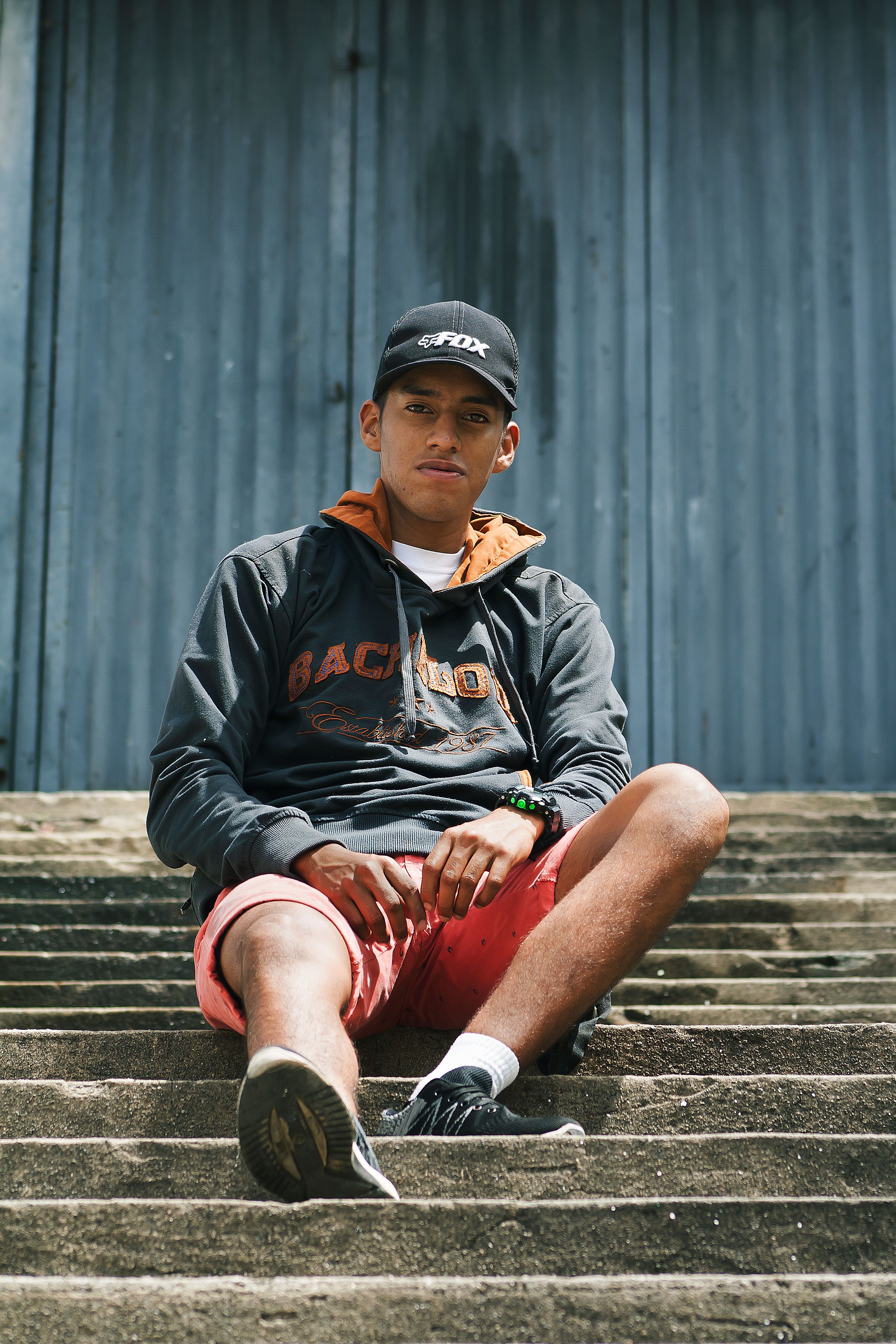 Youthful man sitting on urban stairs, wearing casual hoodie and cap, embodying modern street style.