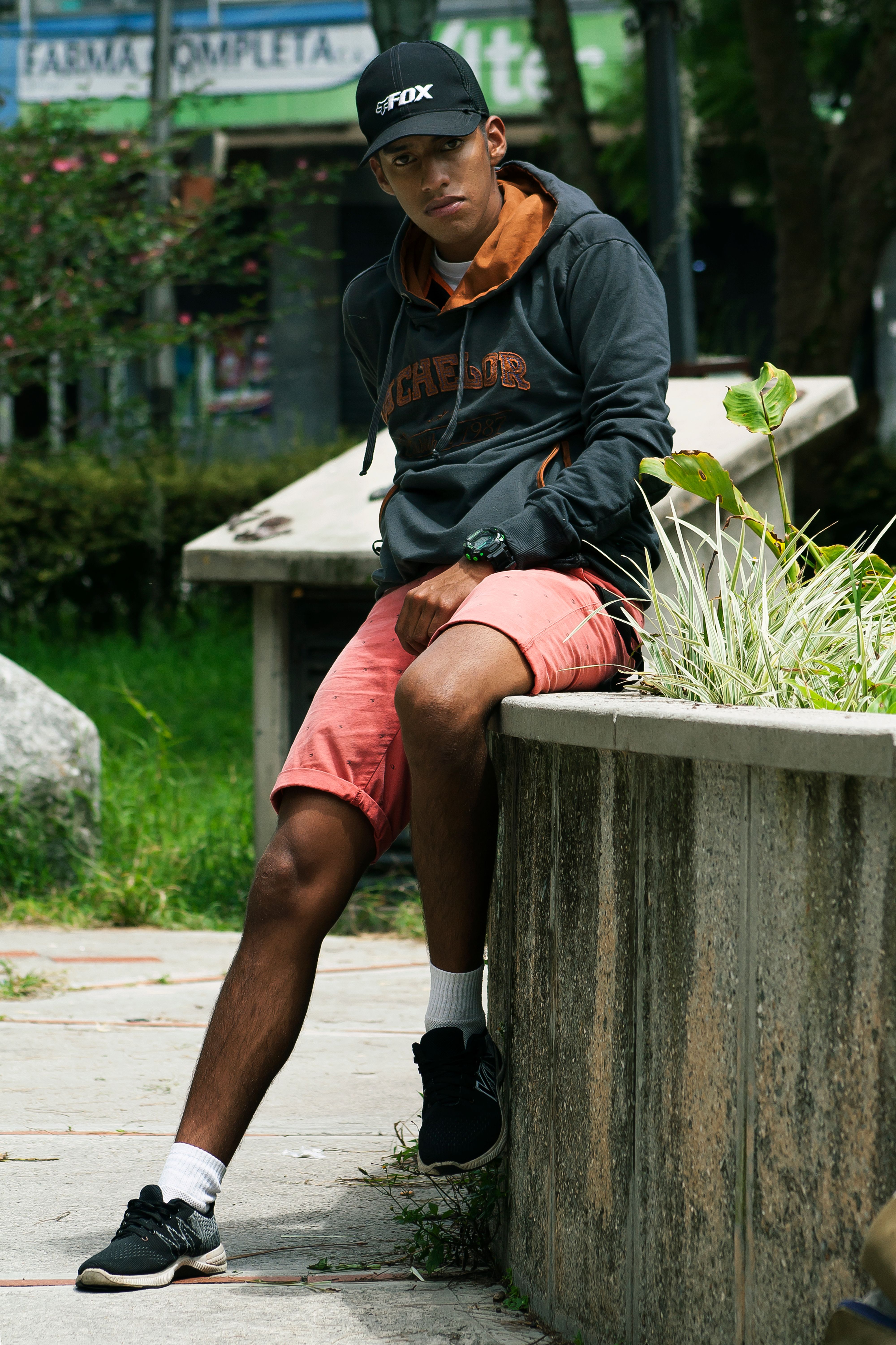 A young man wearing a cap and hoodie sits outdoors on a concrete ledge in a vibrant urban park.