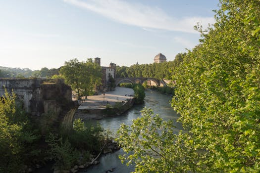Picturesque view of old bridge and lush riverbank in Rome under clear summer sky.