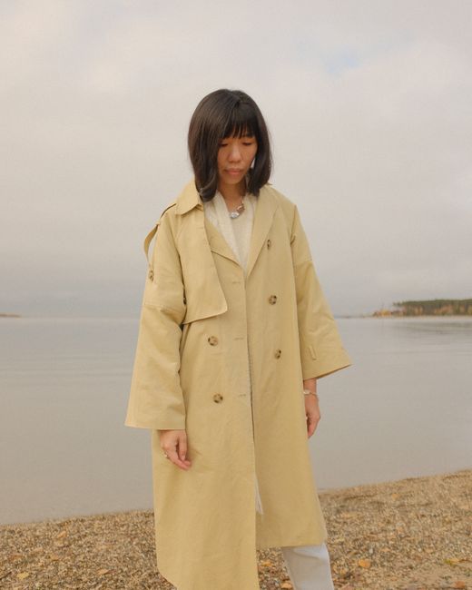 Woman in a trench coat standing by a calm beach during an overcast day.