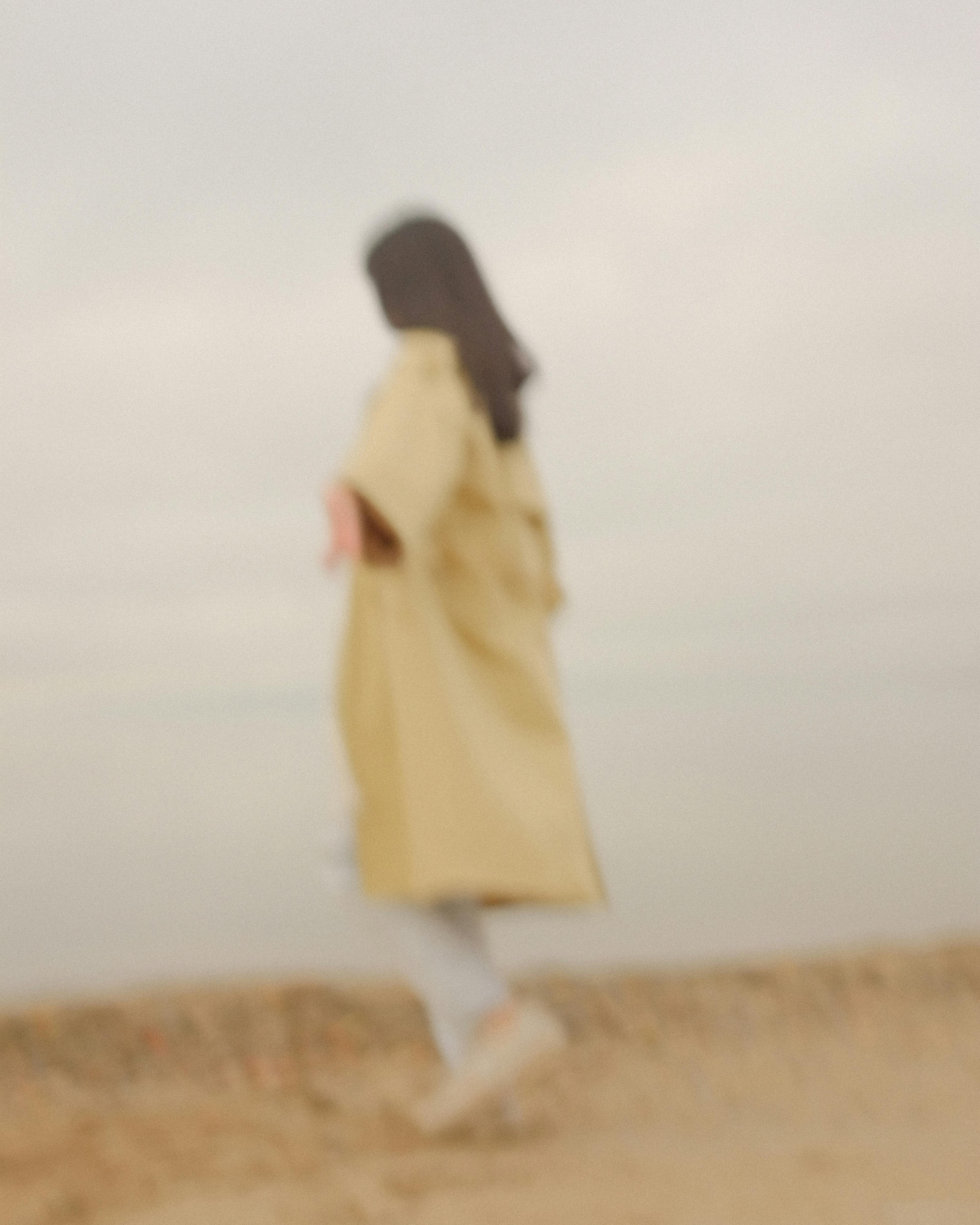 Blurred image of a woman in a coat walking along the beach with a muted color scheme.