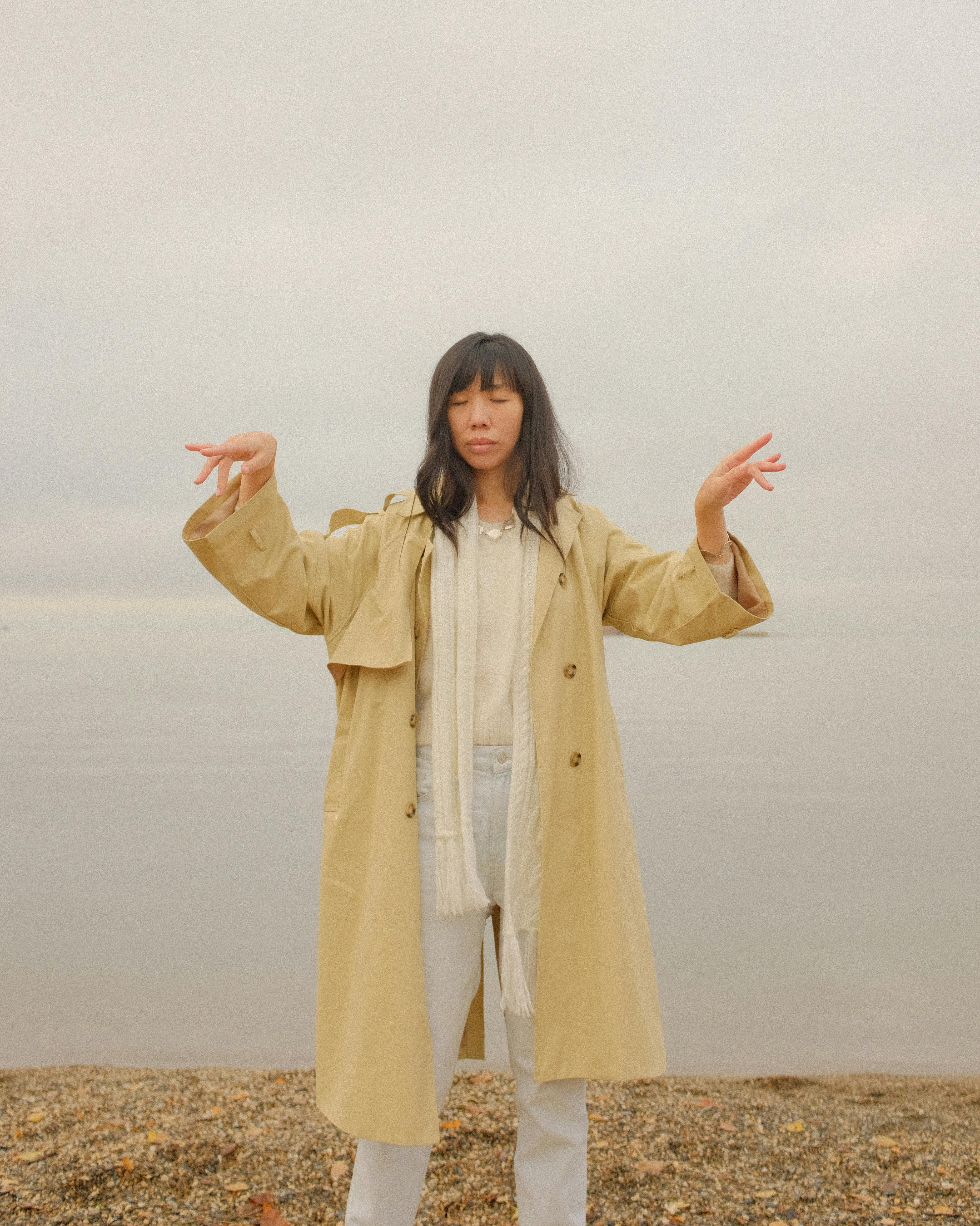 Asian woman in a trench coat with arms raised at a tranquil shore on an overcast day.