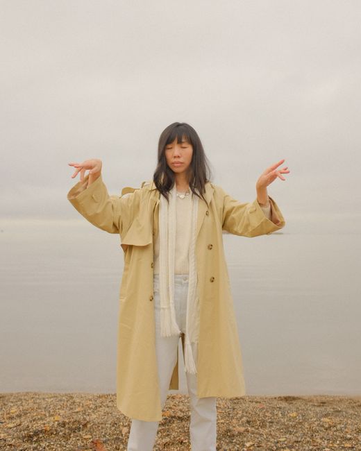 Asian woman in a trench coat with arms raised at a tranquil shore on an overcast day.