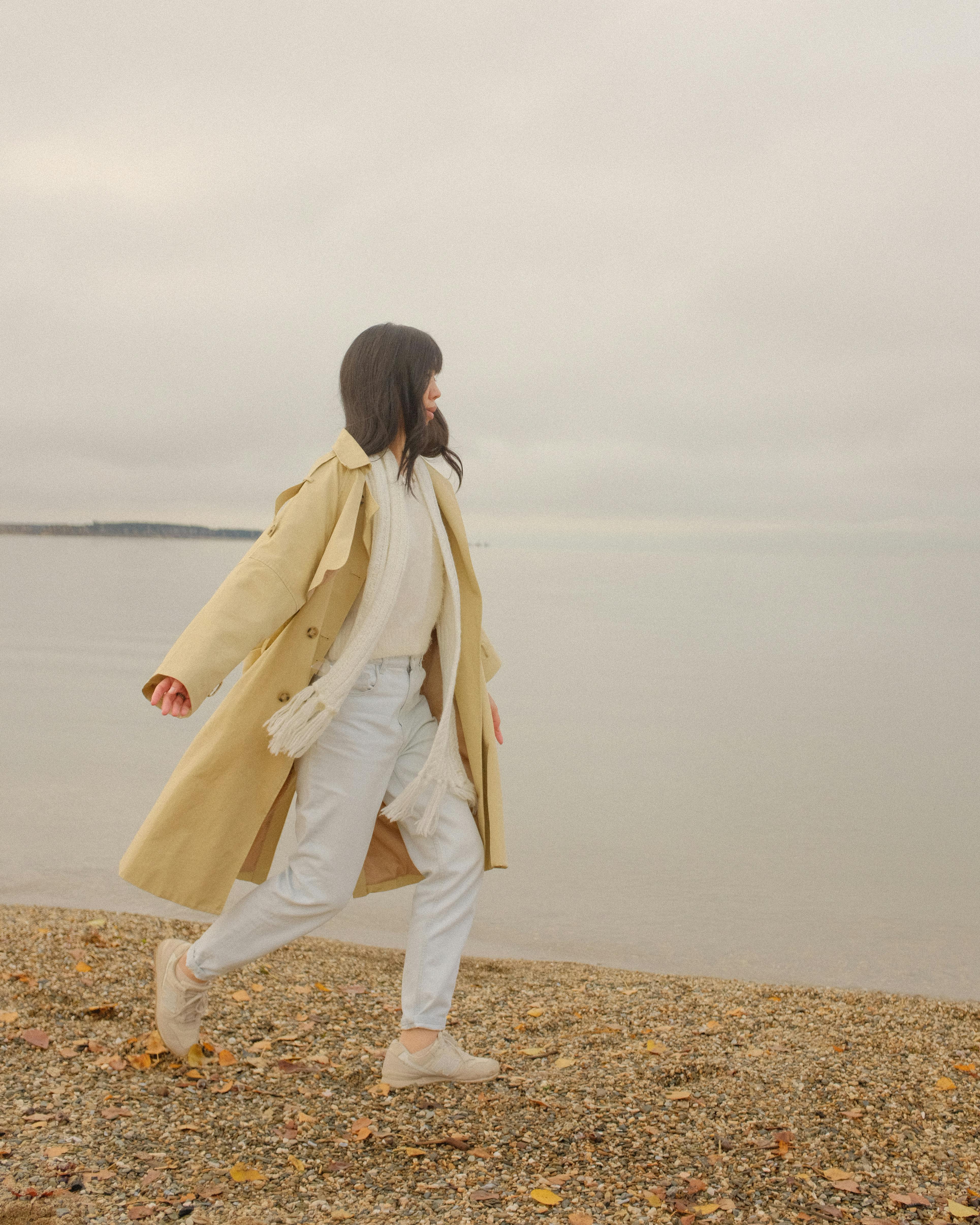 A woman in a beige coat walks along a serene beach, capturing a peaceful autumn day.