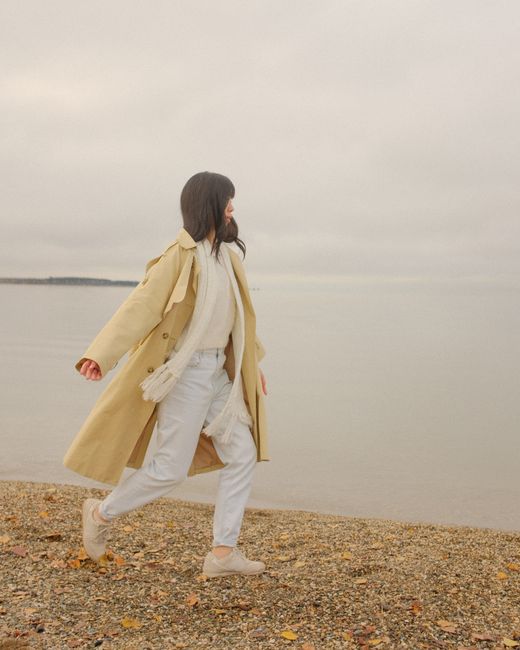 A woman in a beige coat walks along a serene beach, capturing a peaceful autumn day.