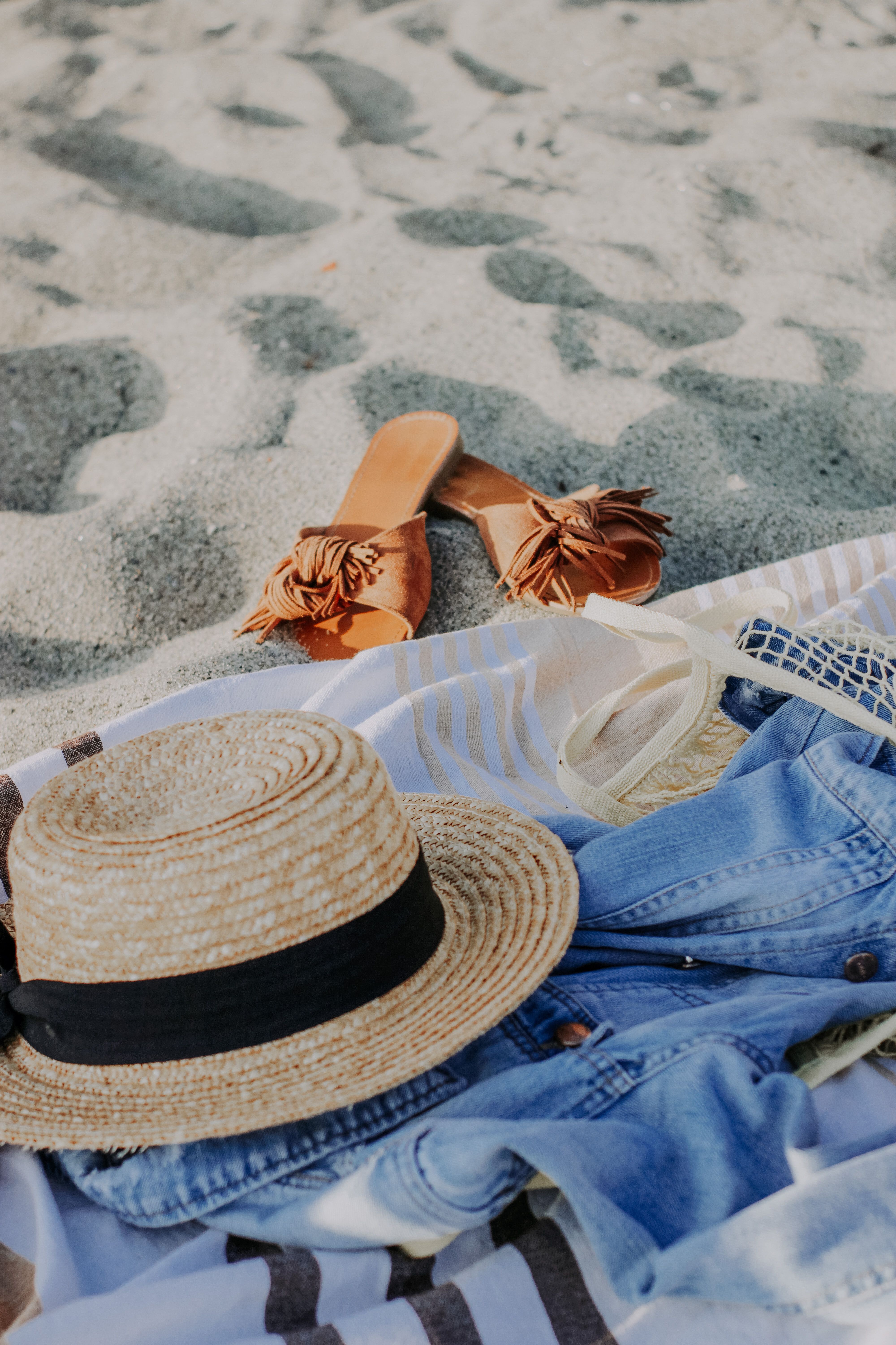 Casual beach essentials including straw hat, denim jacket, and slippers on sand. Perfect for a sunny beach outing.