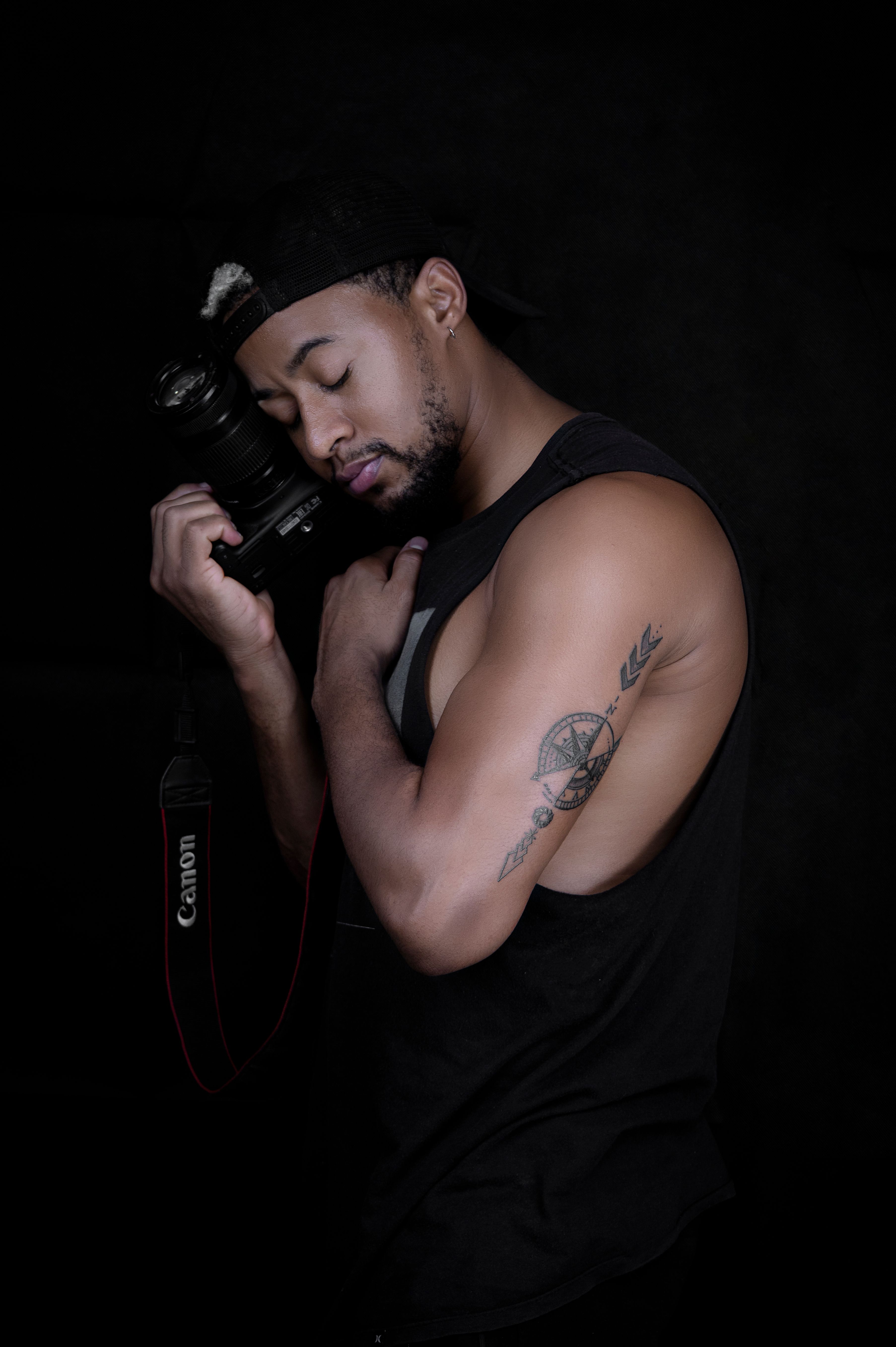 Dark studio portrait of a tattooed man holding a camera with eyes closed.