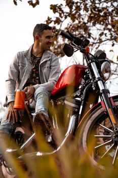 Young man relaxing on a red motorcycle during a fall day outdoors, smiling with a drink in hand.