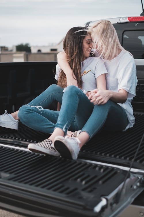 A Same Sex Couple in Denim Jeans and White Shirt Sitting Together on the Car Trunk
