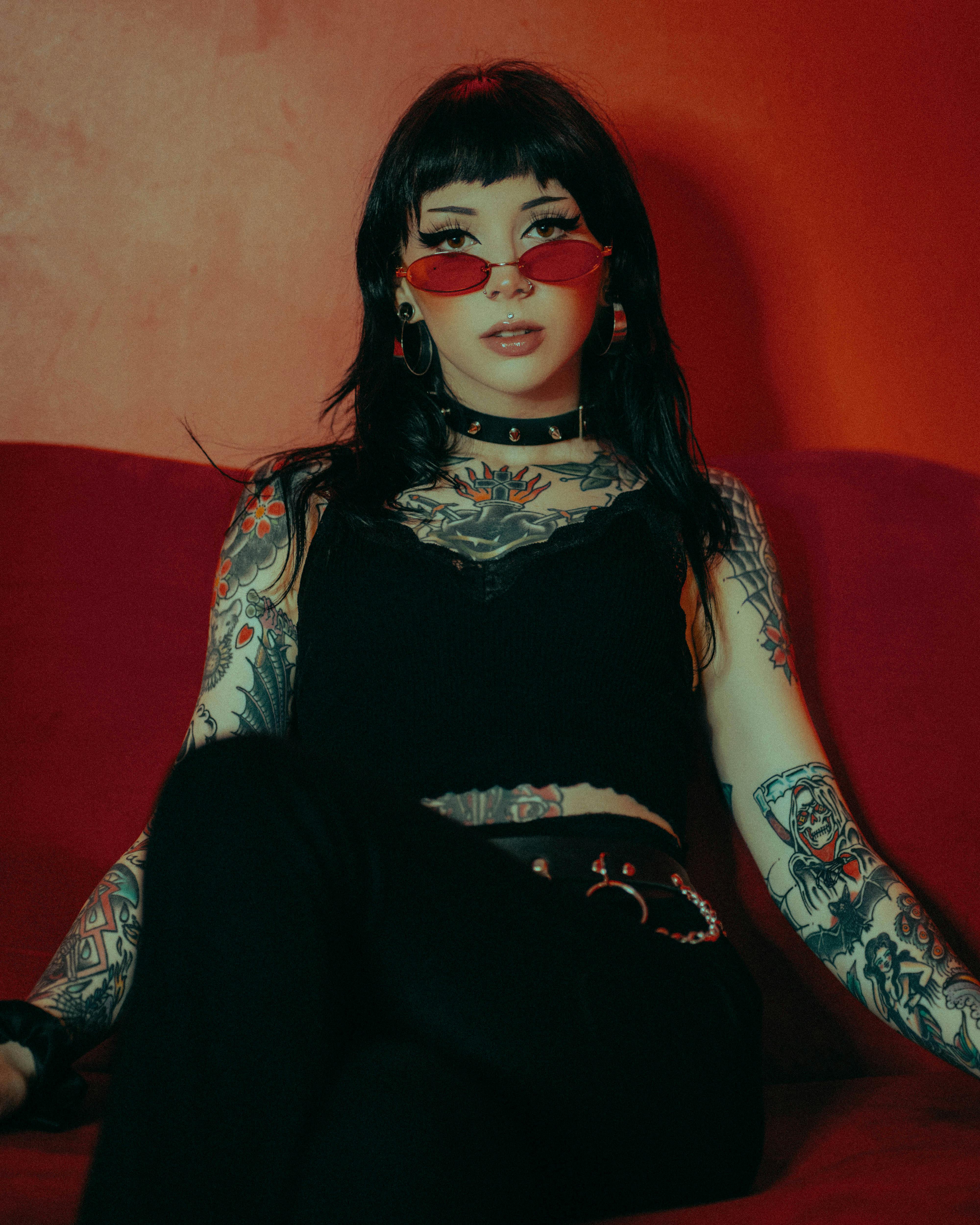 Stylish tattooed woman sitting indoors wearing red sunglasses and black attire.