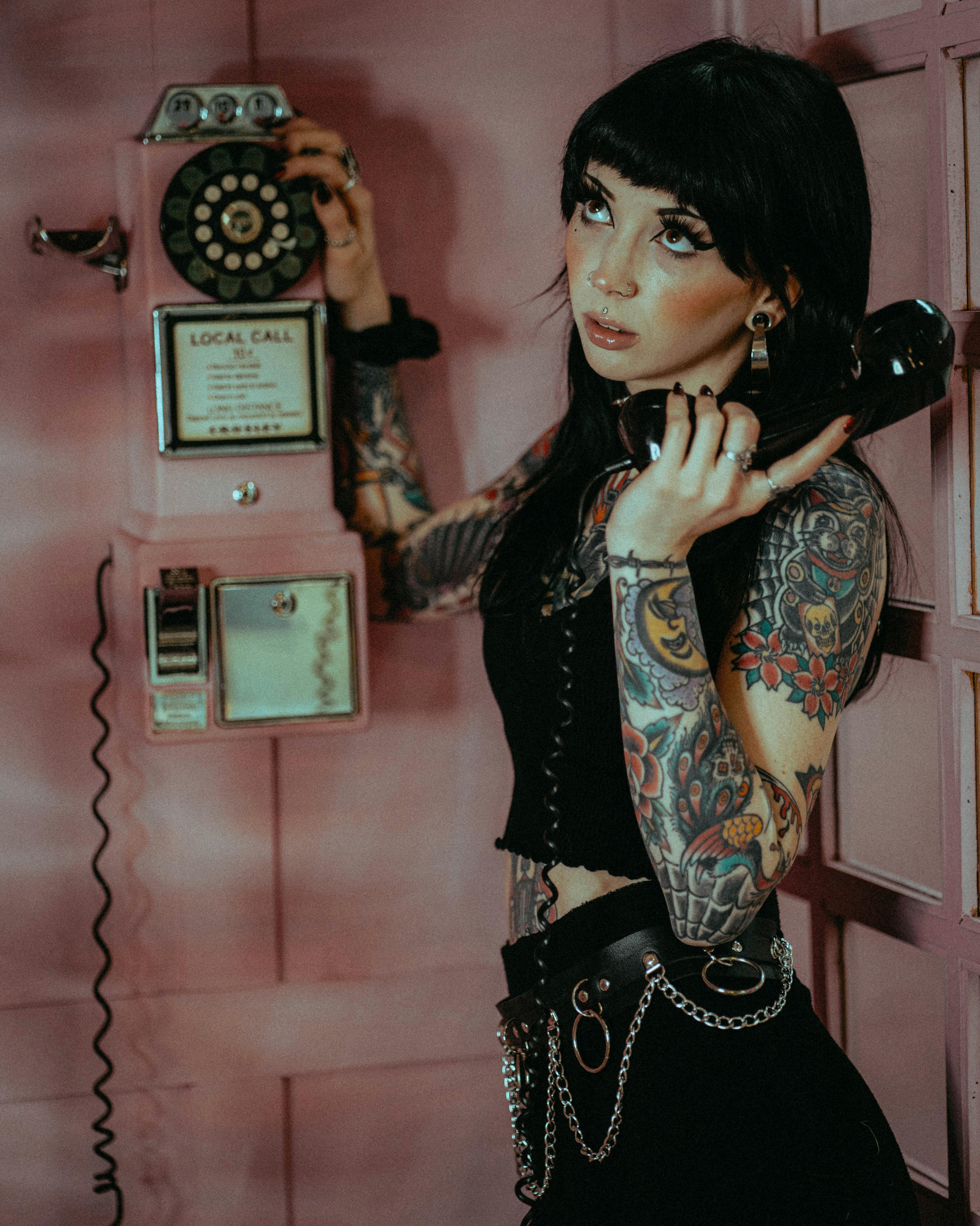 Tattooed woman in black tank top using vintage rotary phone in pink room.