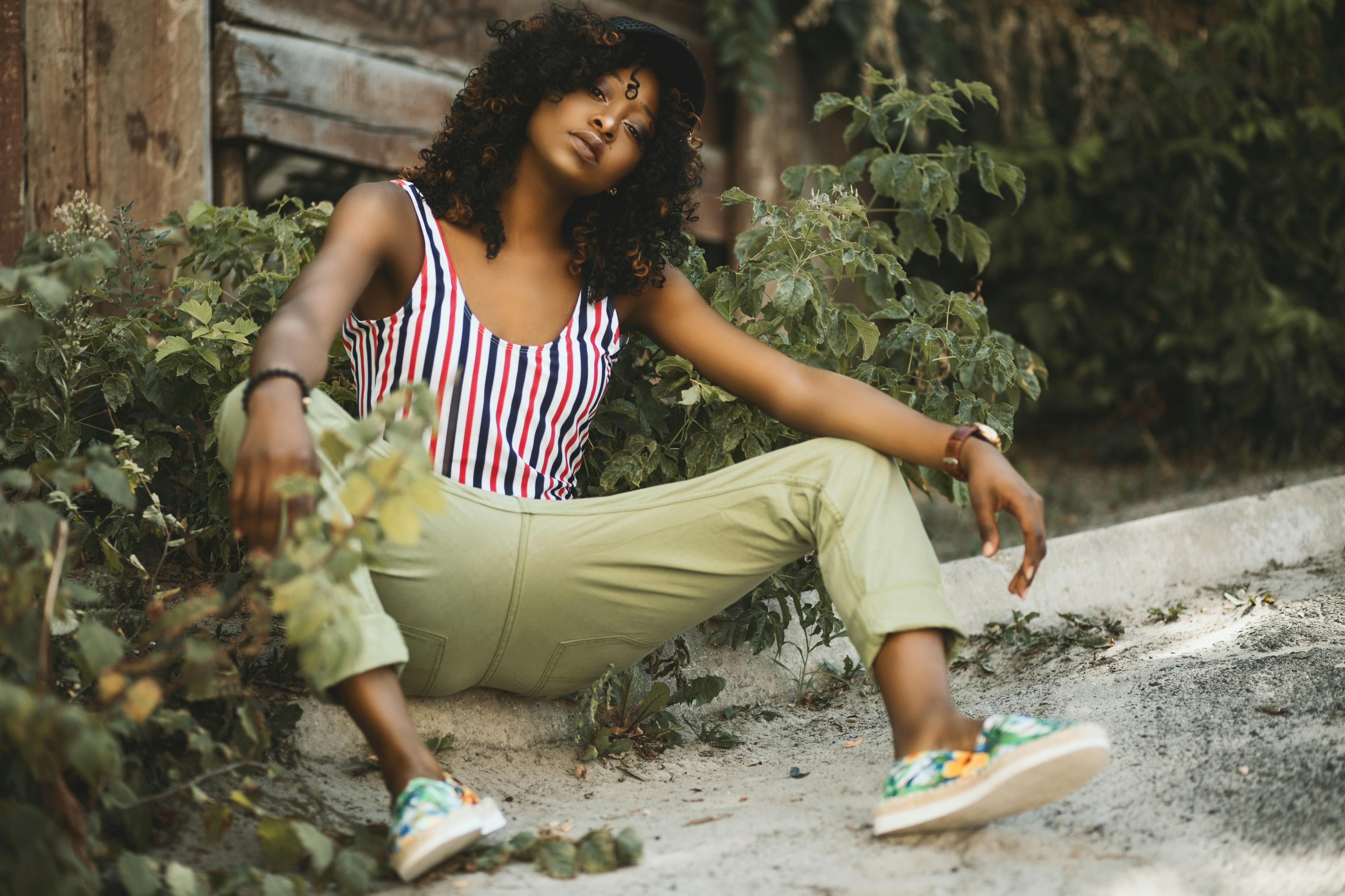 Fashionable young woman with curly hair poses casually in an outdoor setting amidst greenery.