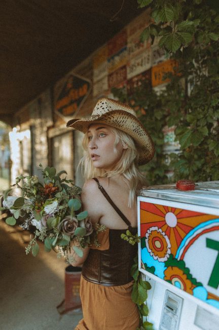 Blonde woman in cowboy hat holding bouquet by vintage decor outside.