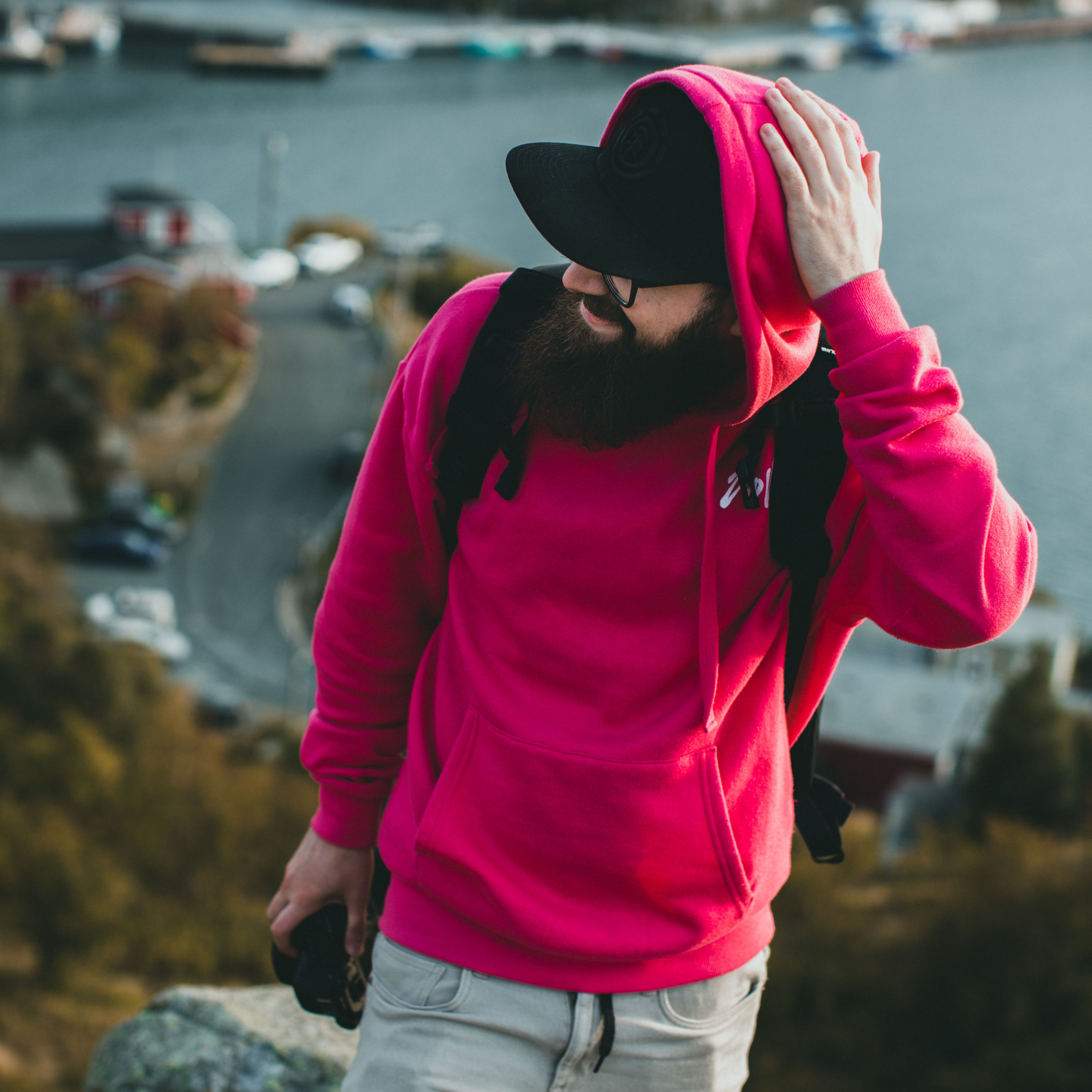 Man with a beard in a bright pink hoodie and black cap enjoying an outdoor landscape.