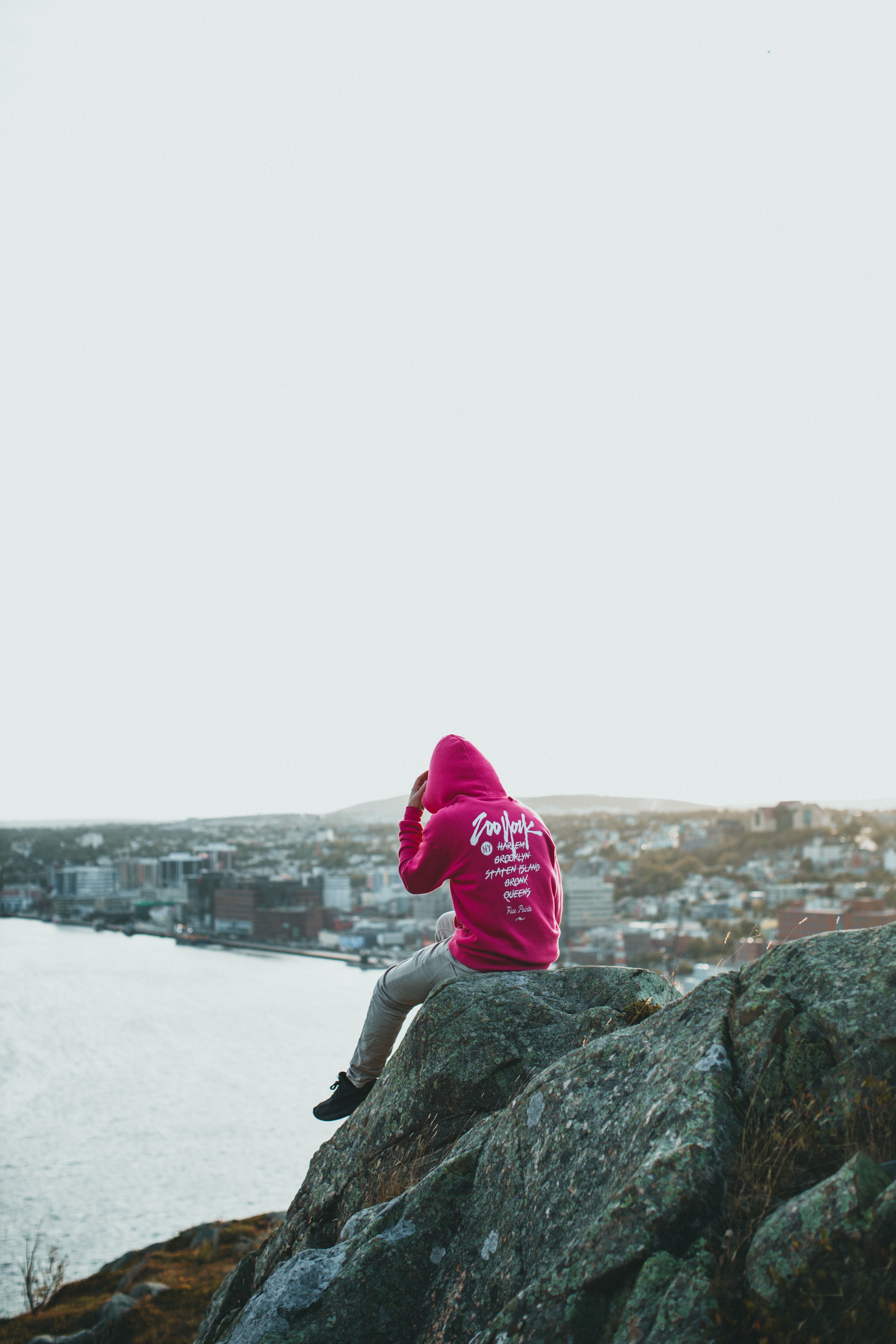 Individual in a pink hoodie sitting on a cliff with a coastal city view, symbolizing adventure and exploration.