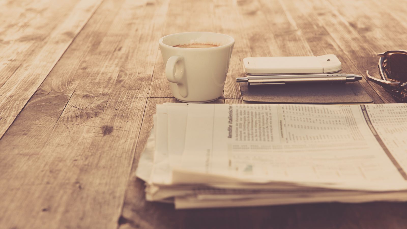 A coffee cup, smartphone, pen and newspaper on a wooden table