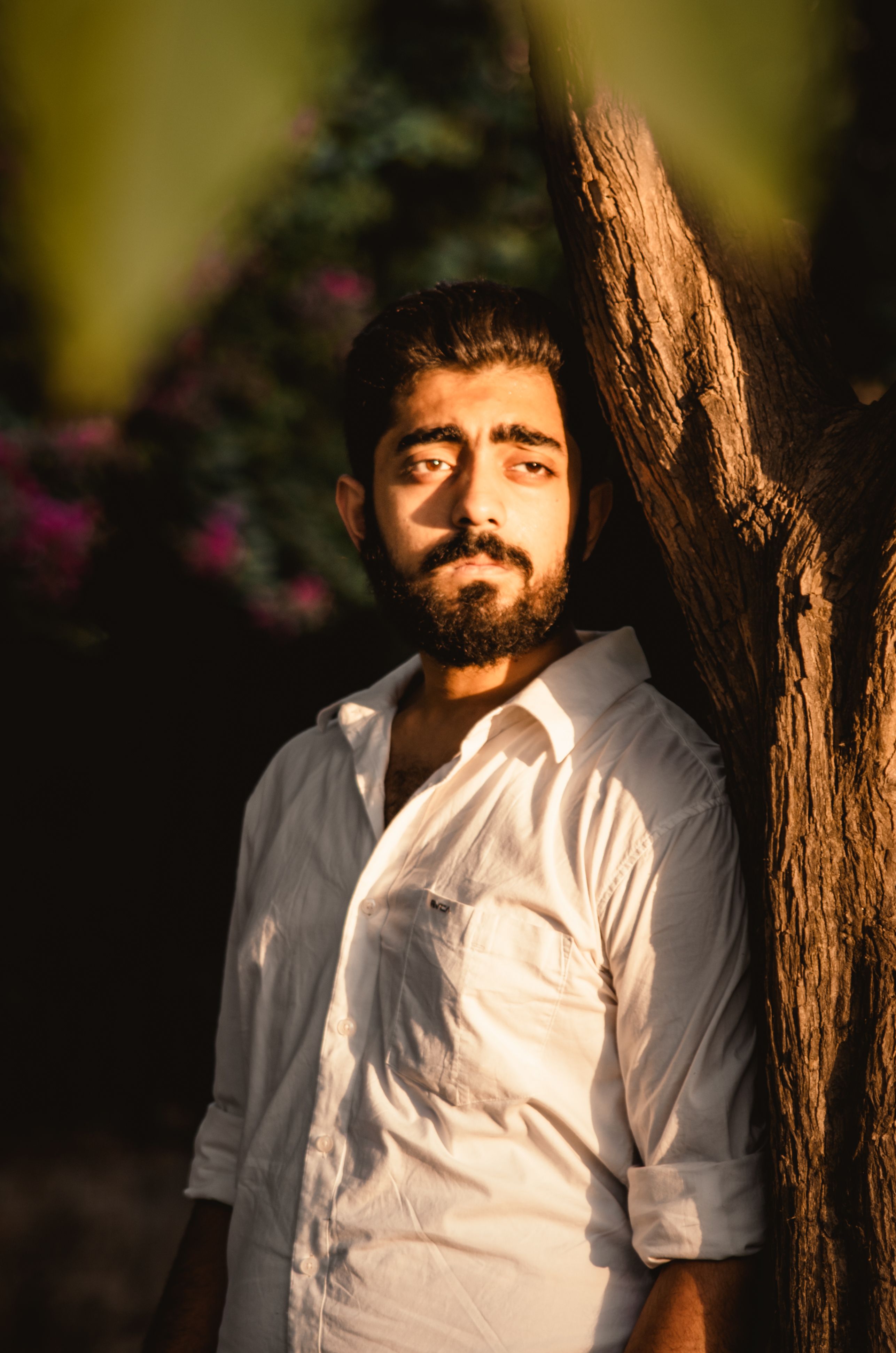 Thoughtful man with beard and white shirt leans on a tree in warm sunlight.