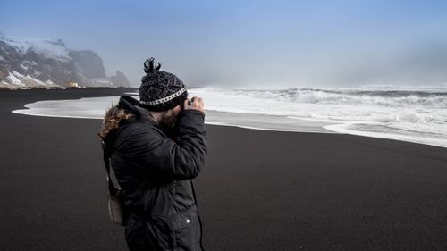 Free A photographer takes pictures on a striking black sand beach with ocean waves on a cloudy day. Stock Photo