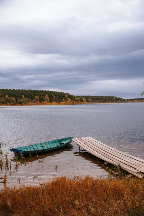 Free Scenic autumn landscape featuring a calm lake with a lone boat and wooden dock, under a cloudy sky. Stock Photo